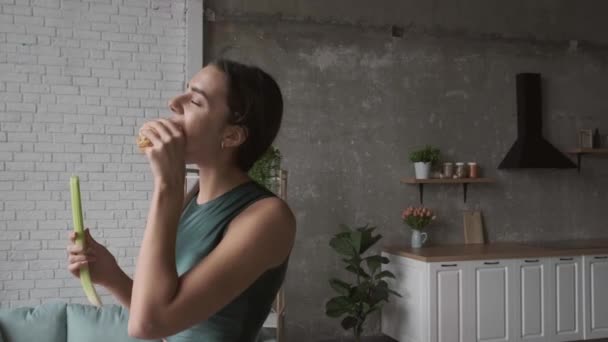 Mulher caucasiana está comendo um hambúrguer enquanto segura um pedaço de alho-porro posando na cozinha usando um vestido — Vídeo de Stock