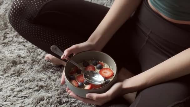 Caucásico en forma mujer está posando en el suelo con las piernas cruzadas sosteniendo un tazón con cereales y frutas — Vídeos de Stock