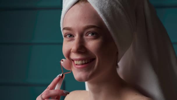 Laughing caucasian woman is massaging her face with a derma roller posing with a towel on head and bare shoulders — Stock Video