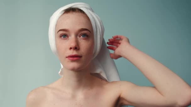 Ginger woman with freckles is posing at camera after a bath wearing a towel on head with bare shoulders on a studio wall — 图库视频影像