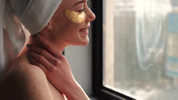 Kaukázusi vörös nő, aki törölközőt és arany hidrogél szemtapaszt visel, csupasz vállakkal mosolyog az ablak előtt. — Stock videók