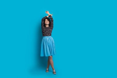 Beyaz kadın stüdyoda vakit geçirip mavi alana yakın poz veriyor. El ele tutuşup elbise giyiyor.