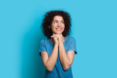 Düşünceli, beyaz, mavi elbiseli, kıvırcık saçlı kadın bir stüdyo duvarında gözlerini kaçıran bir şeyin hayalini kuruyor.