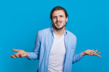 Kararsız adam, mavi bir stüdyonun duvarındaki kameraya düşünceli bir poz vererek avuç içiyle bir şeyleri karşılaştırıyor.