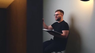 Sakallı ve gözlüklü beyaz bir adam elinde bir kitapla beyaz bir stüdyo duvarında online toplantı yapıyor ve bir şey anlatıyor.