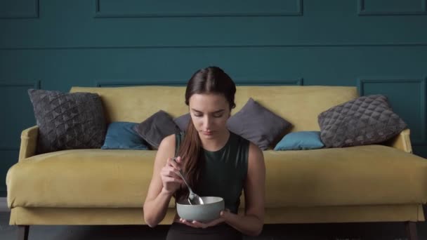 Sportliche Frau in Sportbekleidung sitzt mit überkreuzten Beinen auf dem Boden und isst Müsli — Stockvideo