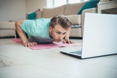 Sarı saçlı beyaz bir adam dizüstü bilgisayar kullanırken yerde şınav çekiyor.