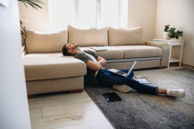 Uykucu Kafkasyalı bir adam bir dizüstü bilgisayarda çalışıyor. Elinde bazı belgeler var. Kanepeye uzanıp dinleniyor.