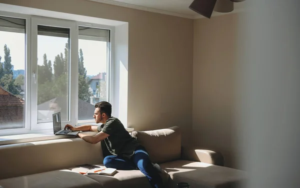 Caucasian freelancer is working remotely with a laptop from home near the window sitting on a couch — Stock Photo, Image