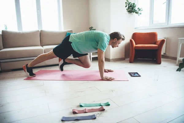 Hombre rubio está usando una tableta mientras hace ejercicios de fitness en casa en una esterilla de yoga — Foto de Stock