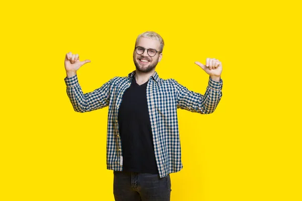 Dumny człowiek gestykuluje na siebie uśmiechając się na żółtej ścianie studia przez okulary noszące brodę — Zdjęcie stockowe