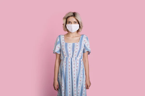 Mulher loira em um vestido de verão longo está usando uma máscara médica em uma parede de estúdio rosa — Fotografia de Stock