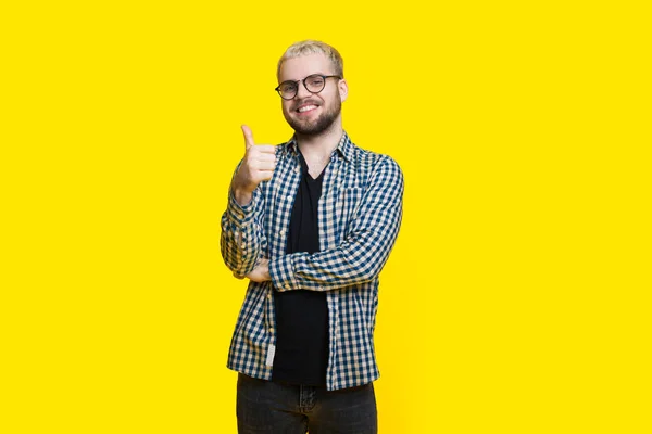 Homem caucasiano loiro com barba e óculos está gesticulando o sinal semelhante com o polegar posando em uma parede de estúdio amarela — Fotografia de Stock