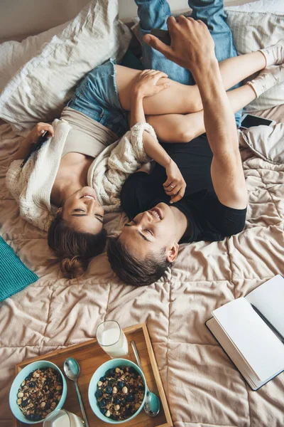 Foto vista superior de um casal caucasiano deitado na cama fazendo uma selfie enquanto lê um livro e come cereais — Fotografia de Stock