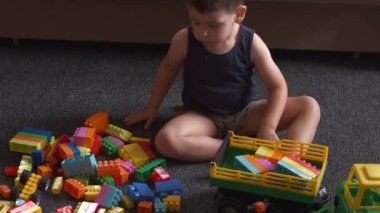 Beyaz çocuk yerde inşaatçıyla oynuyor. Renkli blokları topluyor. 
