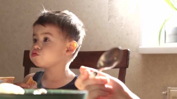 Menino adorável está comendo biscoitos na mesa com seus pais enquanto olha para algo na sala — Vídeo de Stock