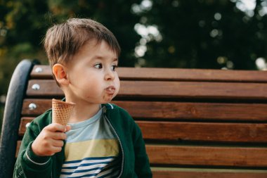 Kafkasyalı çocuk dondurma yiyor. Parktaki bankta oturuyor ve boş alana bakıyor.