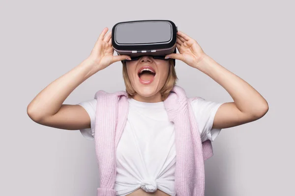 Mujer sonriente con cabello rubio se sorprende por algo mientras usa un auricular de realidad virtual — Foto de Stock