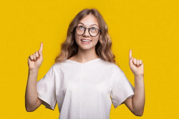 Blonde woman is advertising something pointing up on a yellow studio wall smiling and wearing glasses — Fotografie, imagine de stoc