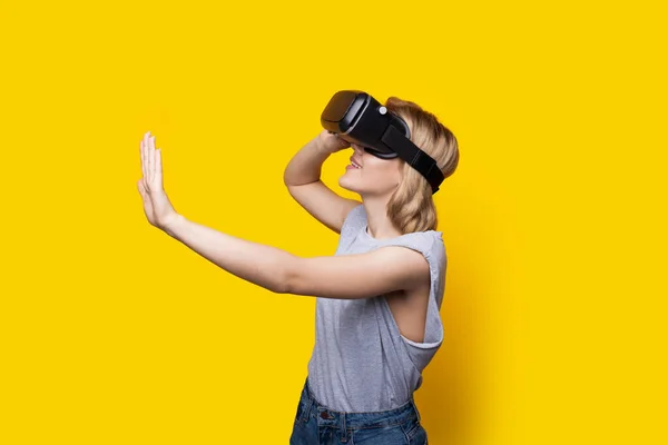 Mujer rubia con auriculares de realidad virtual está tocando algo en una pared de estudio amarillo — Foto de Stock