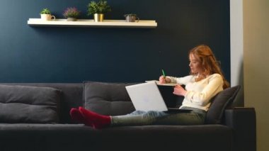 Kızıl kadın kanepede uzanıyor ve deftere yazı yazarken dizüstü bilgisayarda çalışıyor.