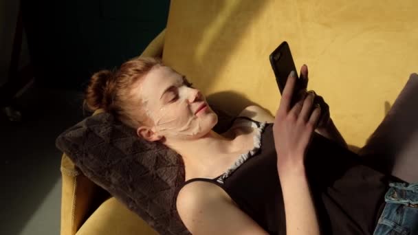 Vista superior de vídeo de una mujer de jengibre caucásica con una máscara de papel mientras charlaba en el móvil y acostado en el sofá — Vídeos de Stock
