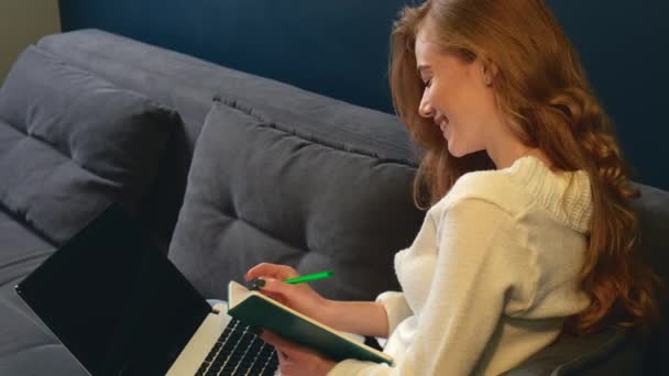 Mujer caucásica está sonriendo mientras hace su tarea en el portátil acostado en el sofá en casa — Vídeo de stock