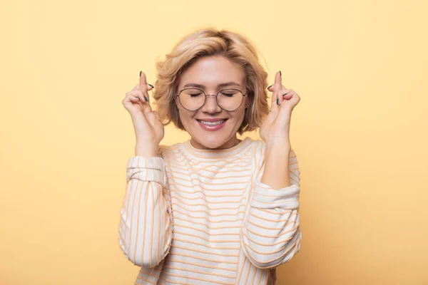 Mulher loira posando em uma parede de estúdio amarelo está sonhando com algo fechando o olho e cruzando os dedos — Fotografia de Stock