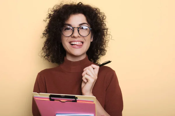 Adorable estudiante con el pelo rizado mostrando su lengua mientras sostiene la carpeta en una pared de estudio amarillo —  Fotos de Stock