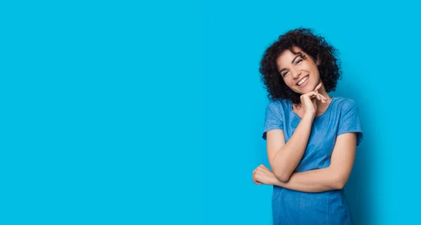 Mujer de pelo rizado está tocando su barbilla y la publicidad de algo en una pared de estudio azul sonriendo a la cámara — Foto de Stock