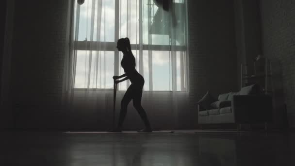 Forma caucásica de la mujer haciendo ejercicios de fitness utilizando una banda cerca de la ventana a una luz baja — Vídeo de stock