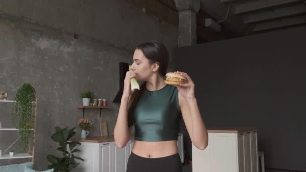 Mujer morena está haciendo la elección entre un puerro y una hamburguesa con ropa deportiva en casa — Vídeos de Stock