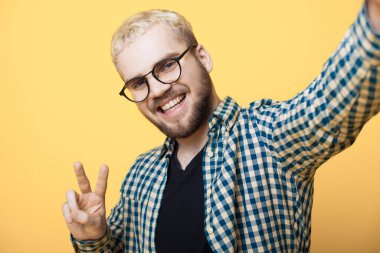 Sakallı ve sarı saçlı beyaz bir adam sarı bir stüdyo duvarında selfie çekiyor. Gülümsüyor ve barış işareti yapıyor.