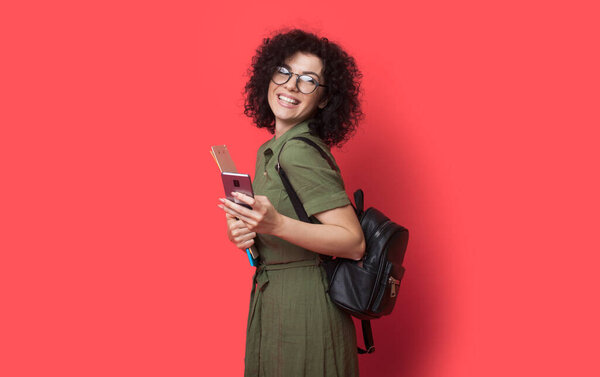 curly haired student with bad is smiling at camera wearing glasses on a red studio wall