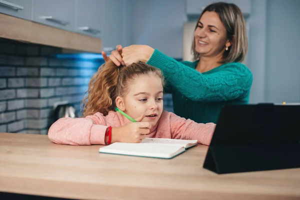 Mãe branca está consertando o cabelo da menina enquanto ela está tendo aulas on-line usando um tablet — Fotografia de Stock