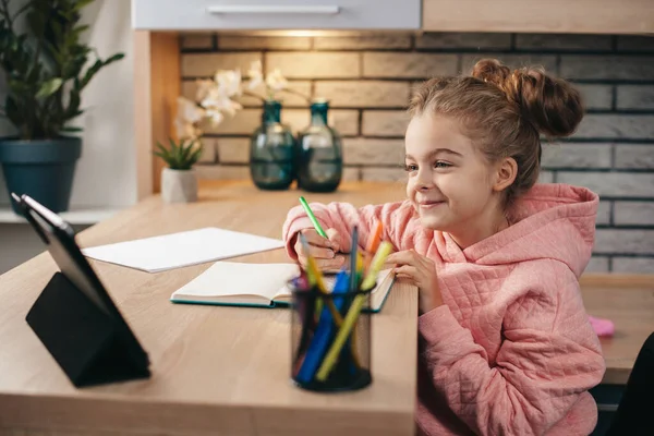 Caucásico linda chica está haciendo su tarea mientras tiene lecciones en línea utilizando una tableta —  Fotos de Stock