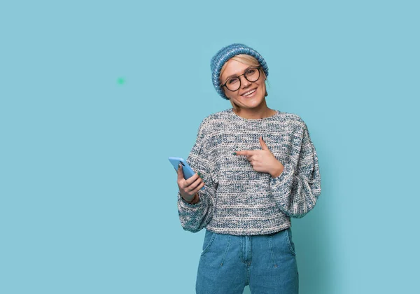 Warm clothed woman is posing on a blue wall pointing at her phone screen smiling at camera