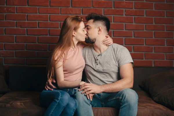 Mulher branca com cabelo vermelho e sardas está sorrindo abraçando seu amante beijando em um sofá em casa — Fotografia de Stock