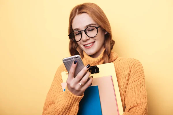Fräknar ingefära student ler på en gul vägg chatta på mobilen och hålla mappar — Stockfoto