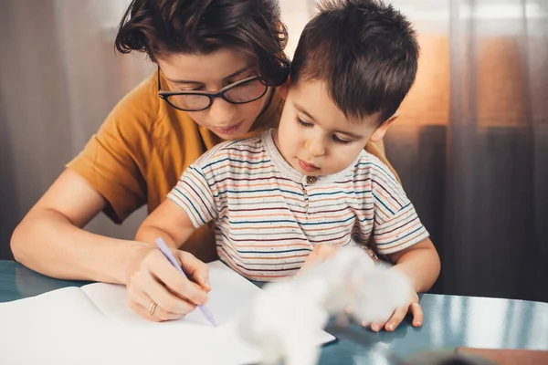 Brunette mother with glasses is helping her son to study and write letters in a copybook