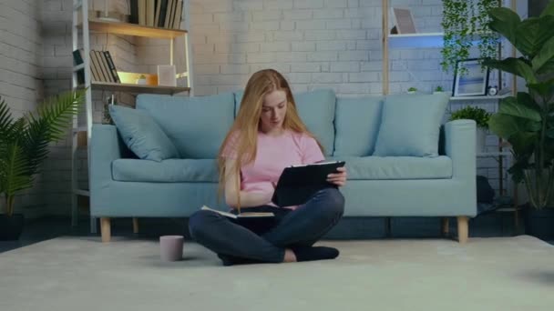 Freckled woman with red hair is having online classes sitting on the floor with a tablet — Stock Video