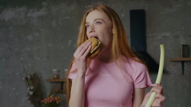 Ginger woman with freckles is eating a burger at home holding a natural leek in other hand — Stock Video