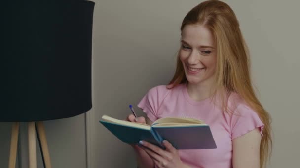 Mujer pelirroja con pecas y cabello rojo está escribiendo algo en un libro y sonríe — Vídeo de stock