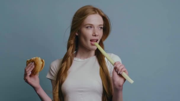 Ginger woman with red hair and freckles at studio biting a leek while holding a hamburger — Stock Video