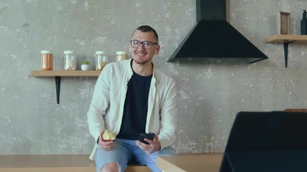 Jovem empresário está tendo uma reunião de vídeo em um laptop enquanto come uma maçã e conversa no celular — Vídeo de Stock