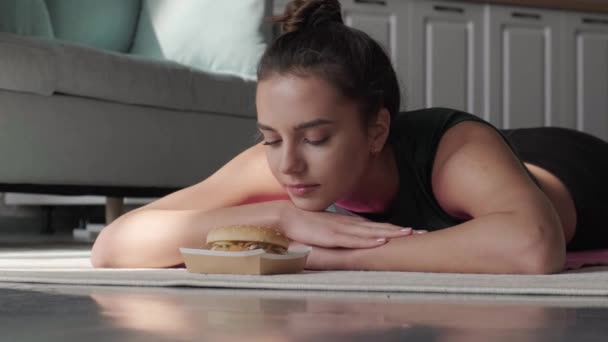 Caucasian woman is lying on the floor near a burger dreaming about eating it — Stock Video