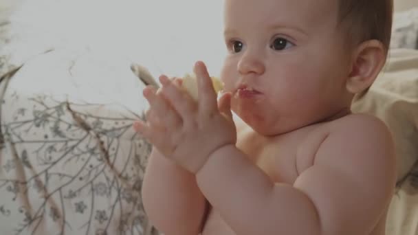 Kleine blanke baby eet een banaan in bed glimlachend naar haar ouders — Stockvideo