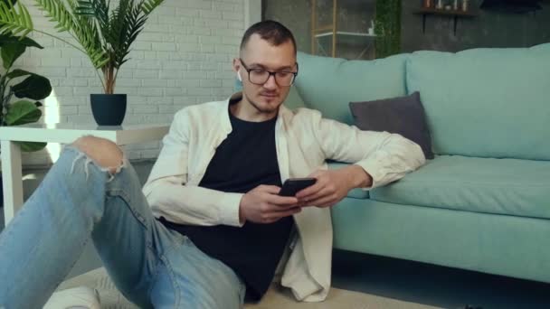 Caucasiano jovem empresário está sentado no chão trabalhando em tablet e usando um telefone perto do sofá — Vídeo de Stock