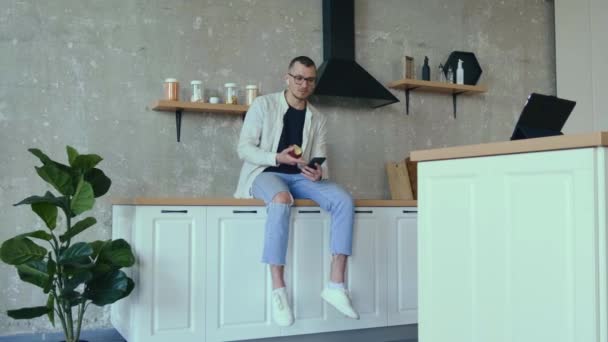 Ung företagare äter ett äpple medan de har ett videomöte i köket med hjälp av en surfplatta — Stockvideo