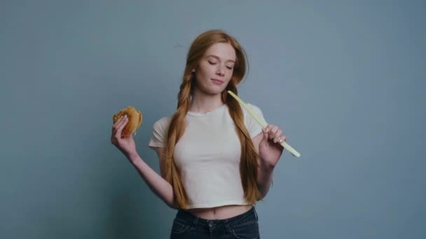 赤い髪とそばかすを持つ白人フィット女性はおいしいハンバーガーと自然なネギの間の難しい決定をしています — ストック動画